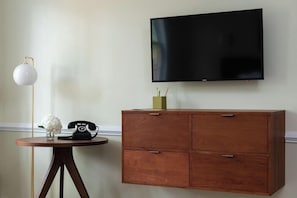 A room with a flat-screen TV mounted on the wall, a wooden cabinet with drawers, a round wooden table with a telephone and a vase of flowers, and a floor lamp.