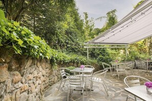 Terrassa exterior amb paviment de pedra, taules i cadires metàl·liques i una pèrgola amb llums penjades.