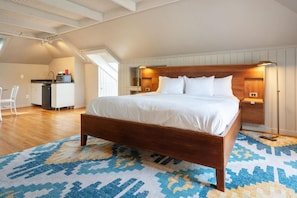 Une chambre Ă coucher avec un lit en bois, du linge de lit blanc, un tapis Ă motifs et, au fond, une petite cuisine.