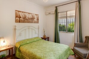 Un dormitorio con cama, una silla, una mesita de noche, una lámpara, una ventana con cortinas y un aire acondicionado instalado en la pared.