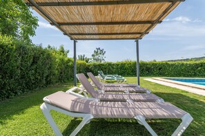 Un área junto a la piscina con sillones de descanso bajo una pérgola sombreada, rodeada de vegetación.