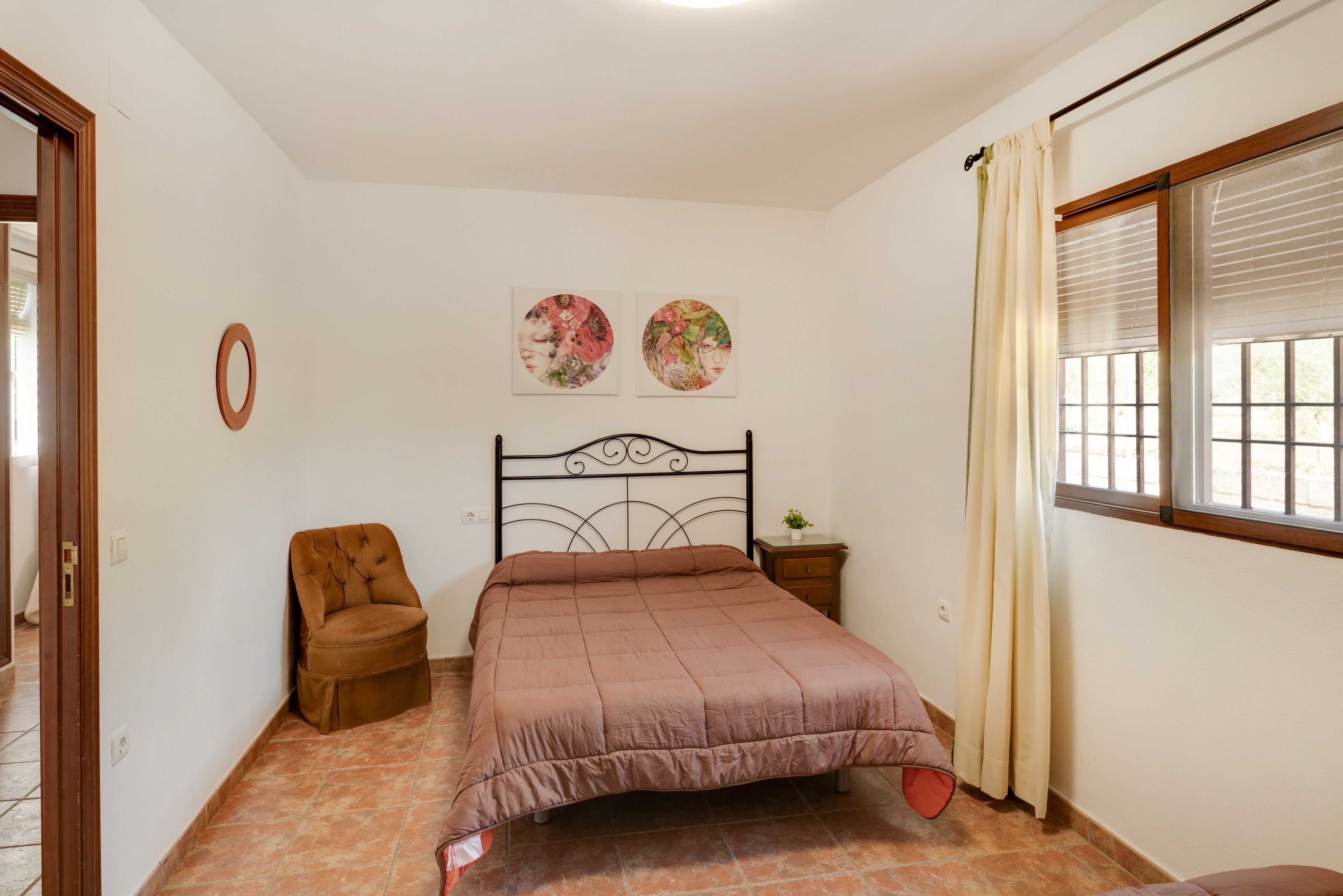 Un dormitorio con cama, una silla, una mesita de noche, una ventana con cortinas y dos cuadros en la pared.