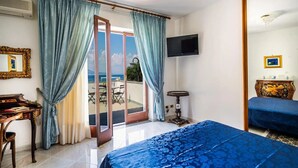 A hotel room with a bed, a desk, a chair, a television, and a view of the sea through a sliding door. - Estate4home - Casa Cinzia (Vico Equense)