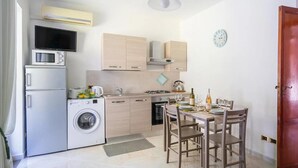 A compact kitchen with a washing machine, microwave, and stove. There is a dining table with chairs and a clock on the wall. - Estate4home - Casa Cinzia (Vico Equense)