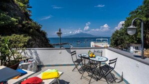 A terrace with a table and chairs, a small table with a tablecloth, a bench with cushions, and a view of the sea with boats. - Estate4home - Casa Cinzia (Vico Equense)