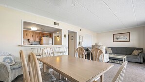 A dining area with a wooden table and chairs, a kitchen in the background, a grey sofa, and a framed picture on the wall.