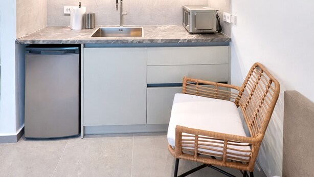 A compact kitchen with a built-in microwave, a sink, and a dish drying rack. A wicker chair is placed in front of the kitchen, and a bed is visible in the foreground. - Villa Lithos Sea View (Paros)
