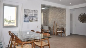 A modern dining area with a glass table and wicker chairs, a stone accent wall, and a view of the outdoors. - Villa Lithos Sea View (Paros)