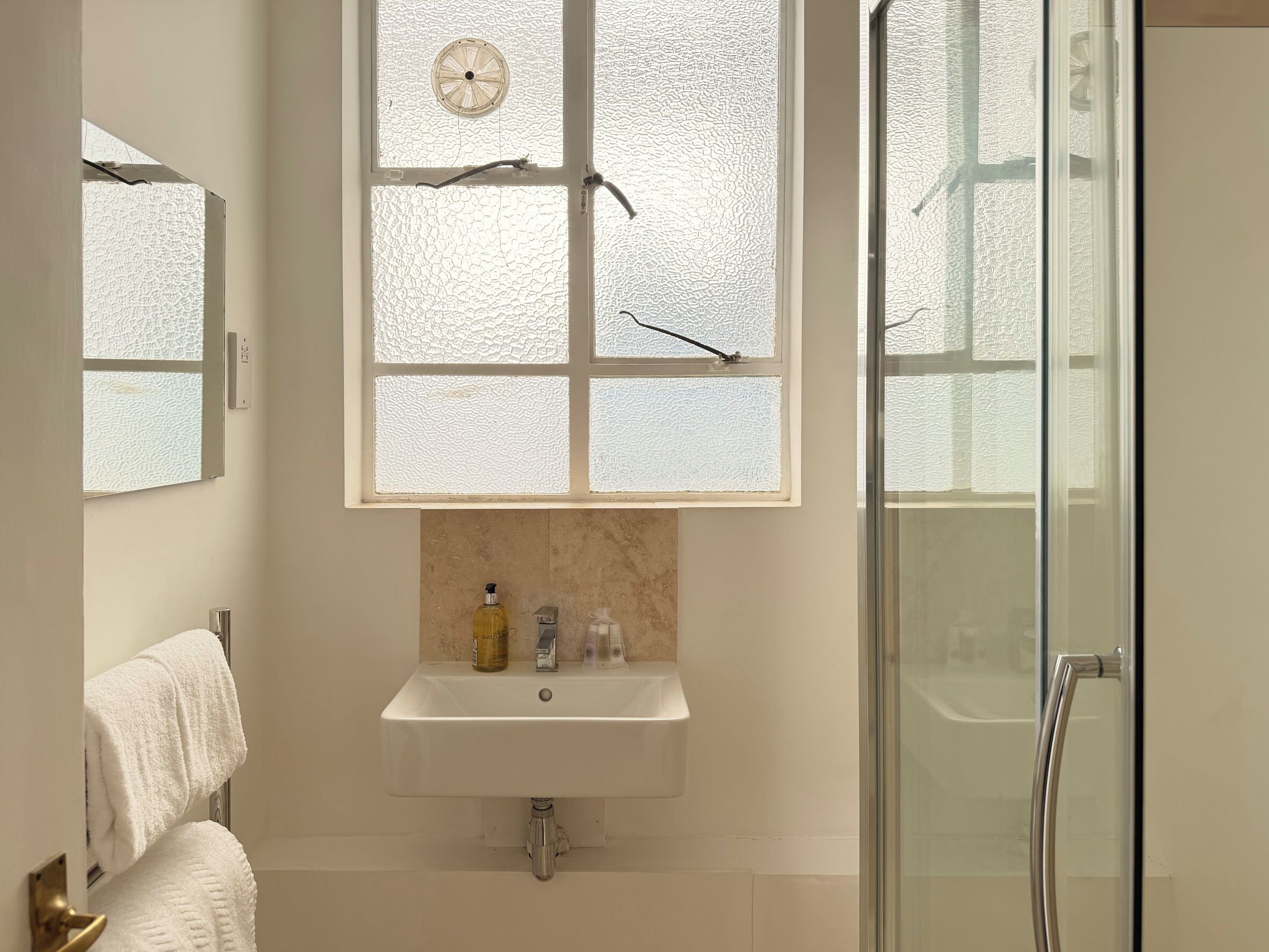 A bathroom with a frosted glass window, a sink with soap dispenser, and a glass shower door.