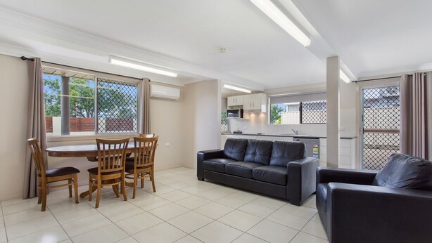 A modern living room with a dining area, a black leather sofa, and a kitchen visible through a glass door. - City Ville Luxury Apartments and Motel (Rockhampton)