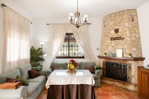 A living room with a stone fireplace, a sofa, a glass-top table with a floral centerpiece, and a chandelier. - Country House 'La Sierrezuela - Families Only' with Mountain Views, Wi-Fi & Air Conditioning (Posadas)