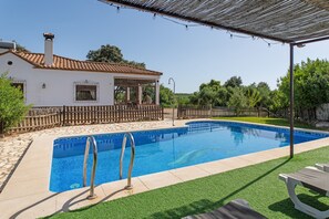 A swimming pool with a wooden deck, a covered patio, and a white house with a tiled roof. - Country House 'La Sierrezuela - Families Only' with Mountain Views, Wi-Fi & Air Conditioning (Posadas)