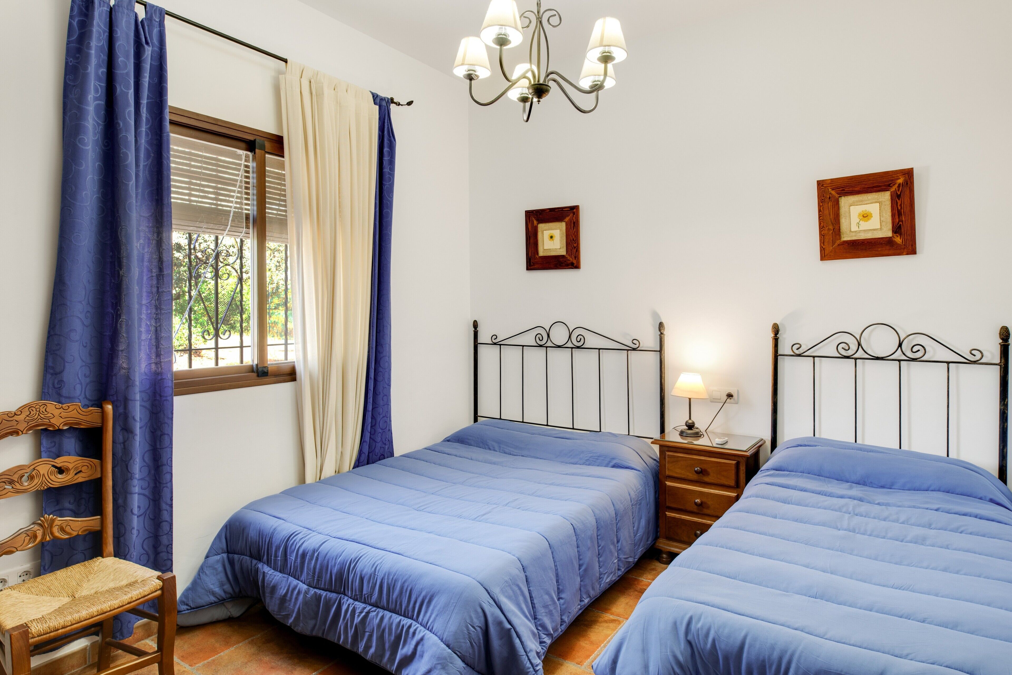 A bedroom with two beds, a wooden chair, a nightstand with a lamp, and a chandelier.