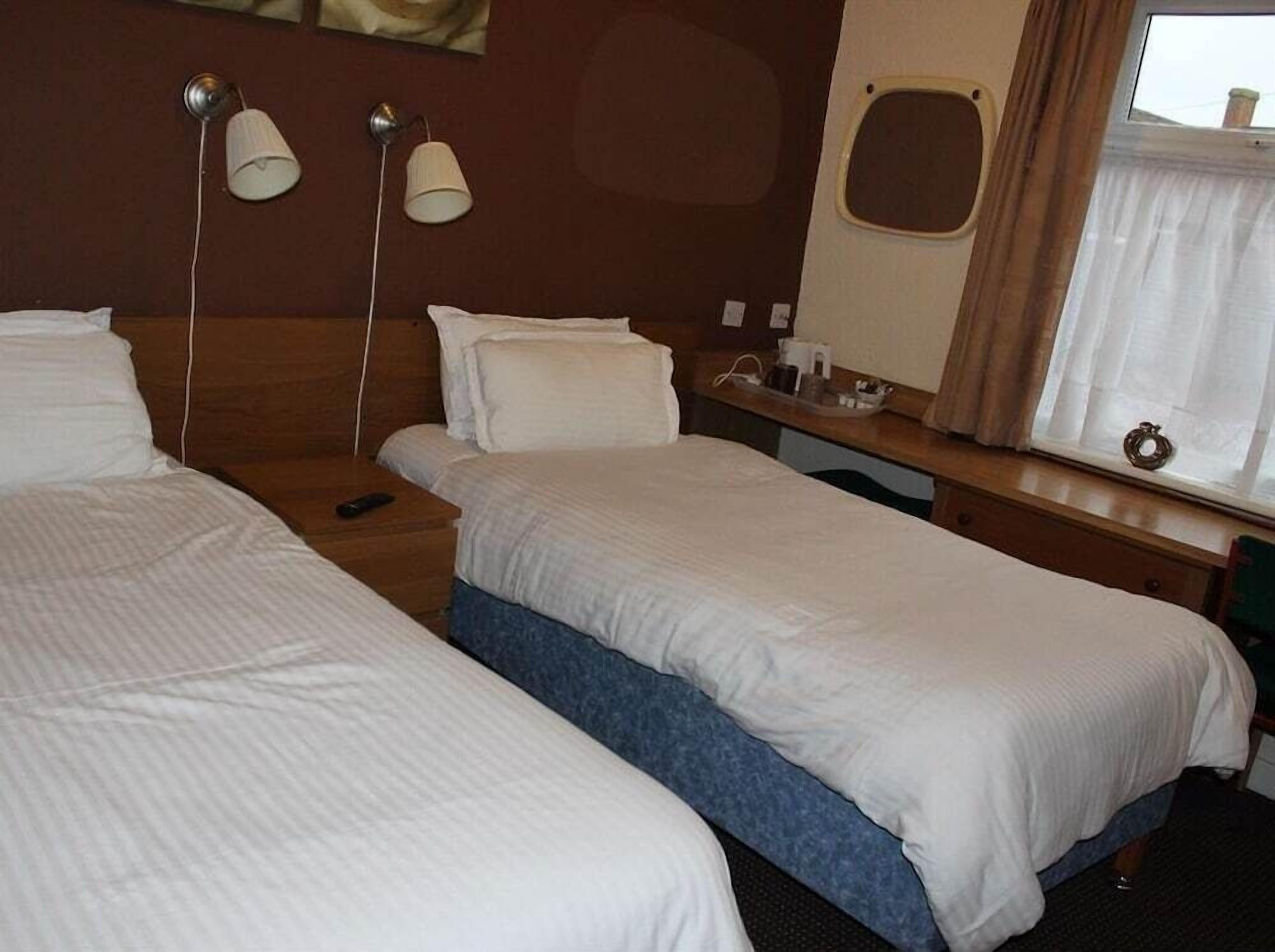 A hotel room with two single beds, a wooden headboard, a bedside table with a lamp, a window with curtains, and a small table with a cup and a phone.