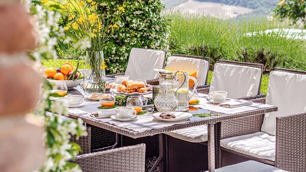 Ein Essbereich im Freien mit Korbstühlen, einem Tisch, der mit Früchten, Gebäck und einer Kanne gedeckt ist.