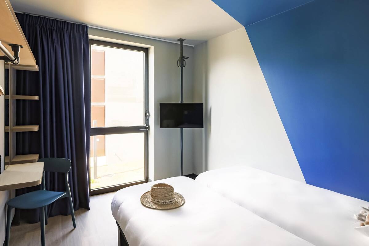 Ein Hotelzimmer mit Bett, Schreibtisch, Stuhl, Fernseher und einem großen Fenster mit Vorhängen.