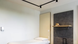 A small, neatly made bed with a white mattress and dark headboard, a wooden floor, a wall-mounted shelf with a hat, and a suitcase.