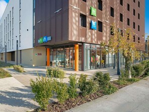 A modern building with a glass entrance, signage for 'ibis' and 'ibis budget', and a well-maintained outdoor area with shrubs and a pathway. - Ibis Budget La Rochelle Centre (La Rochelle)