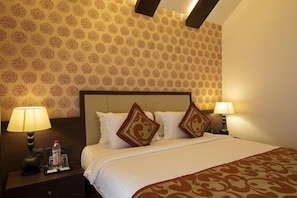 Ein Hotelzimmer mit gemustertem Wandteppich, einem großen Bett mit weißer Bettwäsche und dekorativen Kissen, zwei Nachttischlampen und einem Nachttisch mit Flasche und Gläsern.