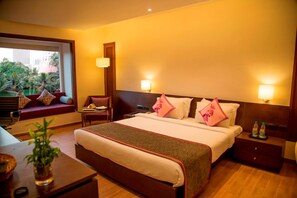 Habitación de hotel con una cama grande, mesitas de noche, un escritorio pequeño, una silla, una ventana con vista a palmeras y una mesita de noche con botellas.