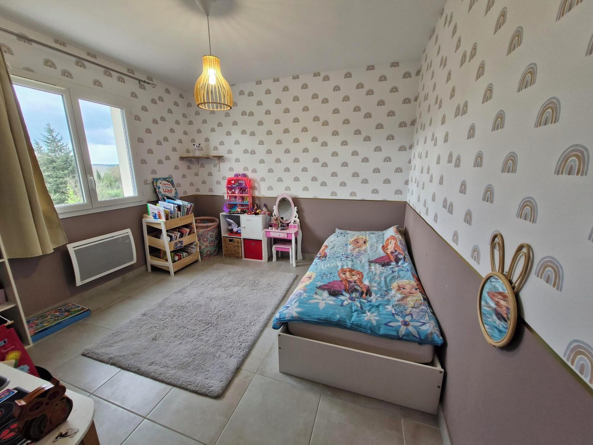 Une chambre d’enfant avec un lit superposé, un bureau, une chaise, un radiateur, une fenêtre et un luminaire suspendu.