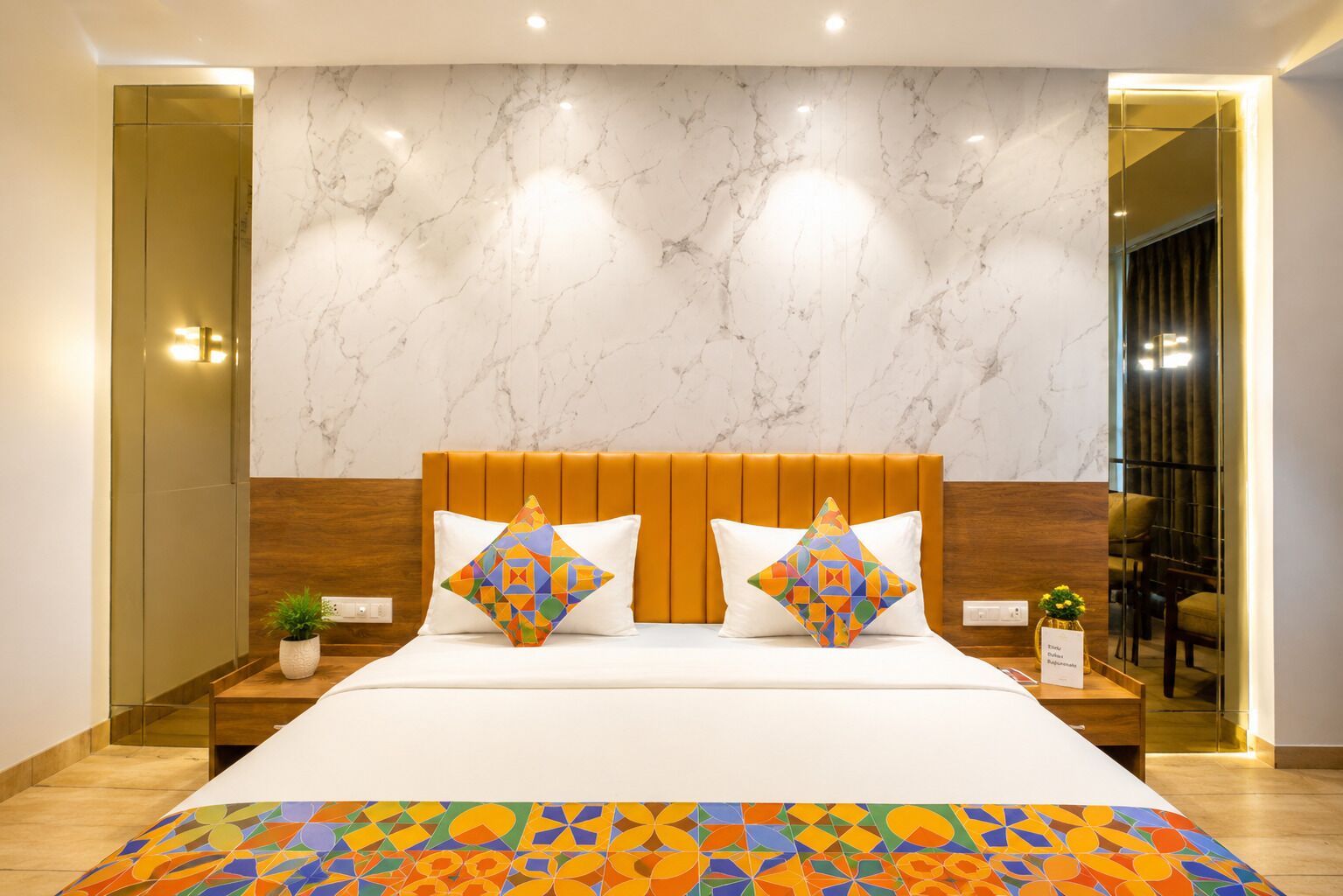 A hotel room with a marble wall, a wooden headboard, a bed with white bedding, two colorful geometric pillows, a small potted plant, and a mirror reflecting the room.