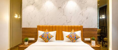 A hotel room with a marble wall, a wooden headboard, a bed with white bedding, two colorful geometric pillows, a small potted plant, and a mirror reflecting the room.