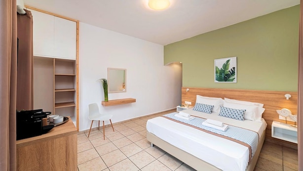 Een moderne hotelkamer met een groot bed, een houten hoofdbord, een klein bureau met een koffieapparaat, een witte stoel en een spiegel.