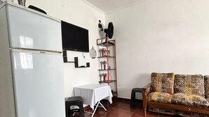 A compact living space with a white refrigerator, a flat-screen TV mounted on the wall, a small table with a white tablecloth, a patterned sofa, and a wooden shelf with decorative items. - Cozy Studio Apartment Praia Grande Ocian (Praia Grande)
