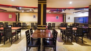 A restaurant with multiple tables and chairs, a central pillar, and a large screen on one wall. - MG Hotel Udayee International (Tirupati)