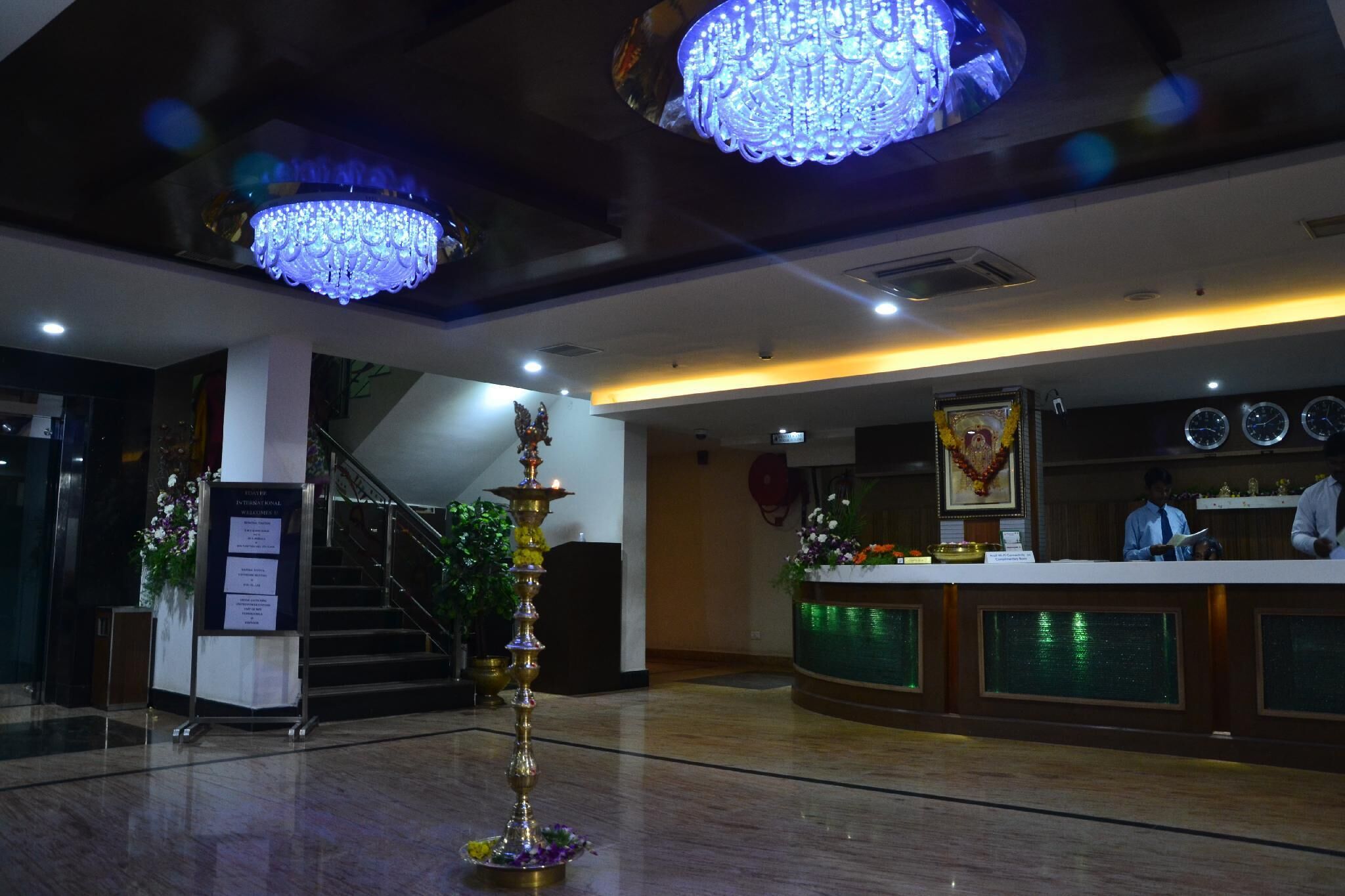 A lobby with a reception desk, a staircase, chandeliers, and a decorative pillar.