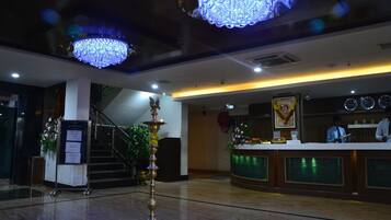 A lobby with a reception desk, a staircase, chandeliers, and a decorative pillar.