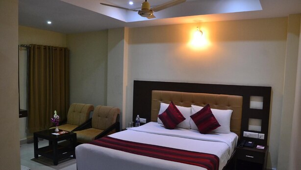 A hotel room with a bed, two armchairs, a nightstand, a bedside table, a ceiling fan, and a wall-mounted light. - MG Hotel Udayee International (Tirupati)