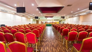 A conference room with rows of red chairs, a wooden floor, and a stage with a screen at the front. - MG Hotel Udayee International (Tirupati)