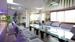 A modern living room with a glass coffee table, grey sofas, a treadmill, and a large window with curtains. - MG Hotel Udayee International (Tirupati)
