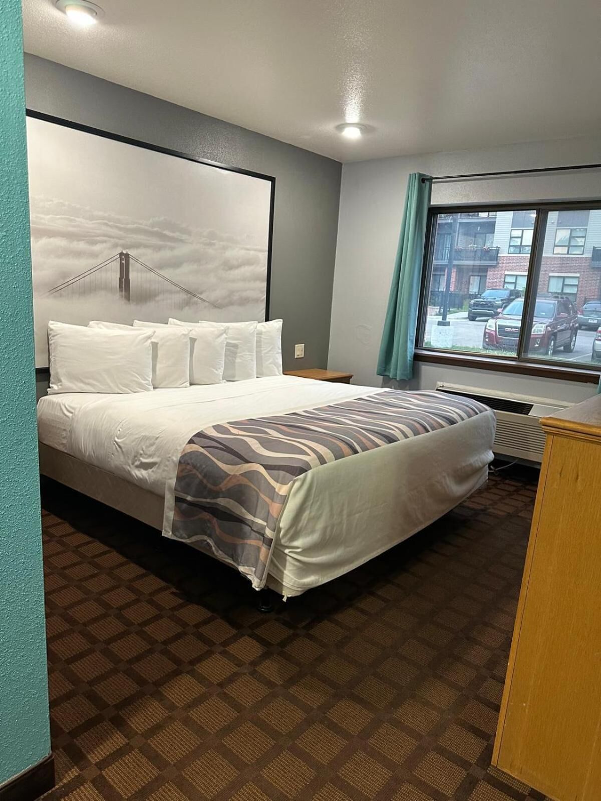 Une chambre d’hôtel avec un lit, une fenêtre donnant sur des immeubles, un mur orné d’un tableau et un luminaire au plafond.