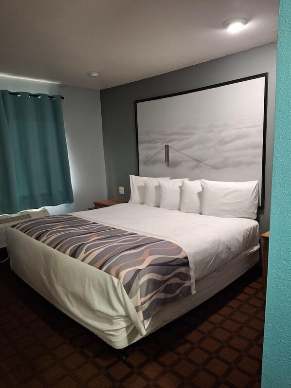 A hotel room with a bed, a headboard, a nightstand, a window with curtains, and a wall-mounted picture. - Mcgoverns Hotel and Suites (Sun Prairie)