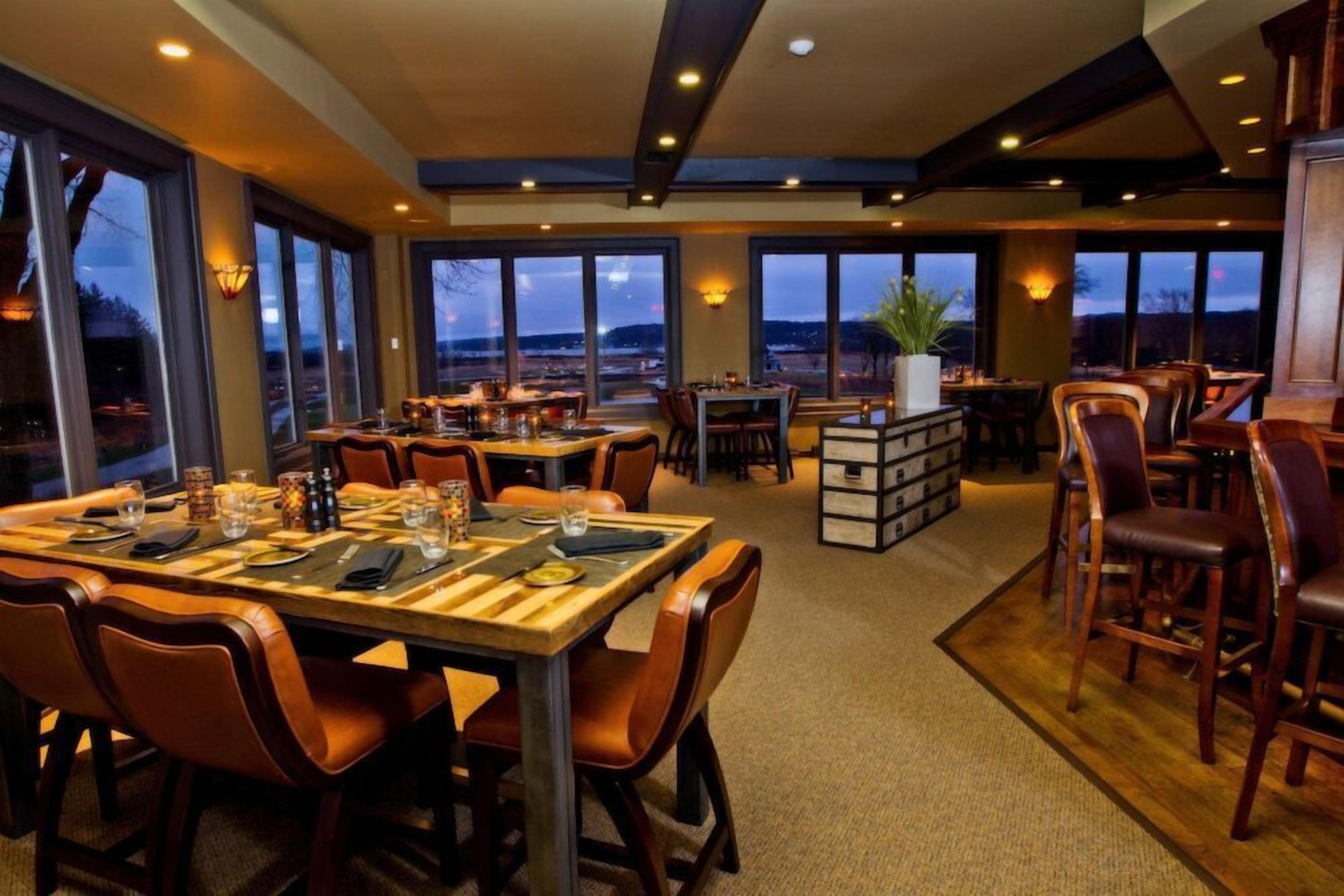 Un restaurante con varias mesas y sillas, ventanas amplias con vistas y una planta en el centro.
