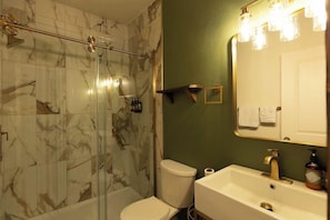 Un bany amb una cabina de dutxa de vidre, un vàter blanc, un lavabo amb un aixeta daurada i un mirall amb il·luminació.