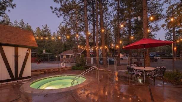 Piscina con sombrilla roja y mobiliario exterior, rodeada de árboles y luces colgantes.