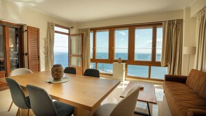 Un coin repas avec une table en bois, des chaises et un vase. De grandes fenêtres offrent une vue sur l’océan.