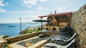 Un espace extérieur aménagé avec des murs en pierre, offrant une vue sur la mer, un navire à l’horizon et un bâtiment en pierre doté d’une cheminée.