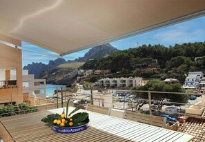 Un balcon donnant sur une ville côtière, une table avec un panier de fruits et une montagne en arrière-plan.