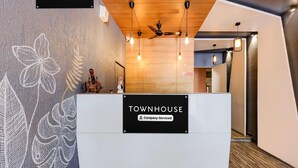 Een receptiebalie met een bord waarop 'TOWNHOUSE Company-Serviced' staat, een plafondventilator, hangende lampen en een houten wand met bladmotieven.