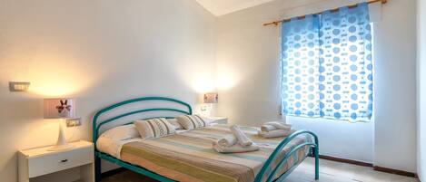Une chambre à coucher comprenant un lit, des tables de chevet, une lampe, une fenêtre avec des rideaux et un luminaire fixé au mur.
