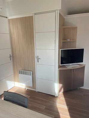 A room with a wardrobe, a TV on a cabinet, and a vent on the wall. - •\tCharming air-conditioned bungalow in a campsite near the sea (Port-la-Nouvelle)