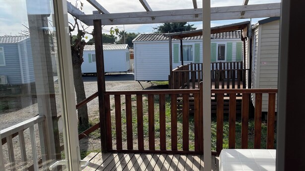 Eine überdachte Terrasse im Freien mit Holzgeländer, einer weißen Bank und Blick auf Mobilheime im Hintergrund.