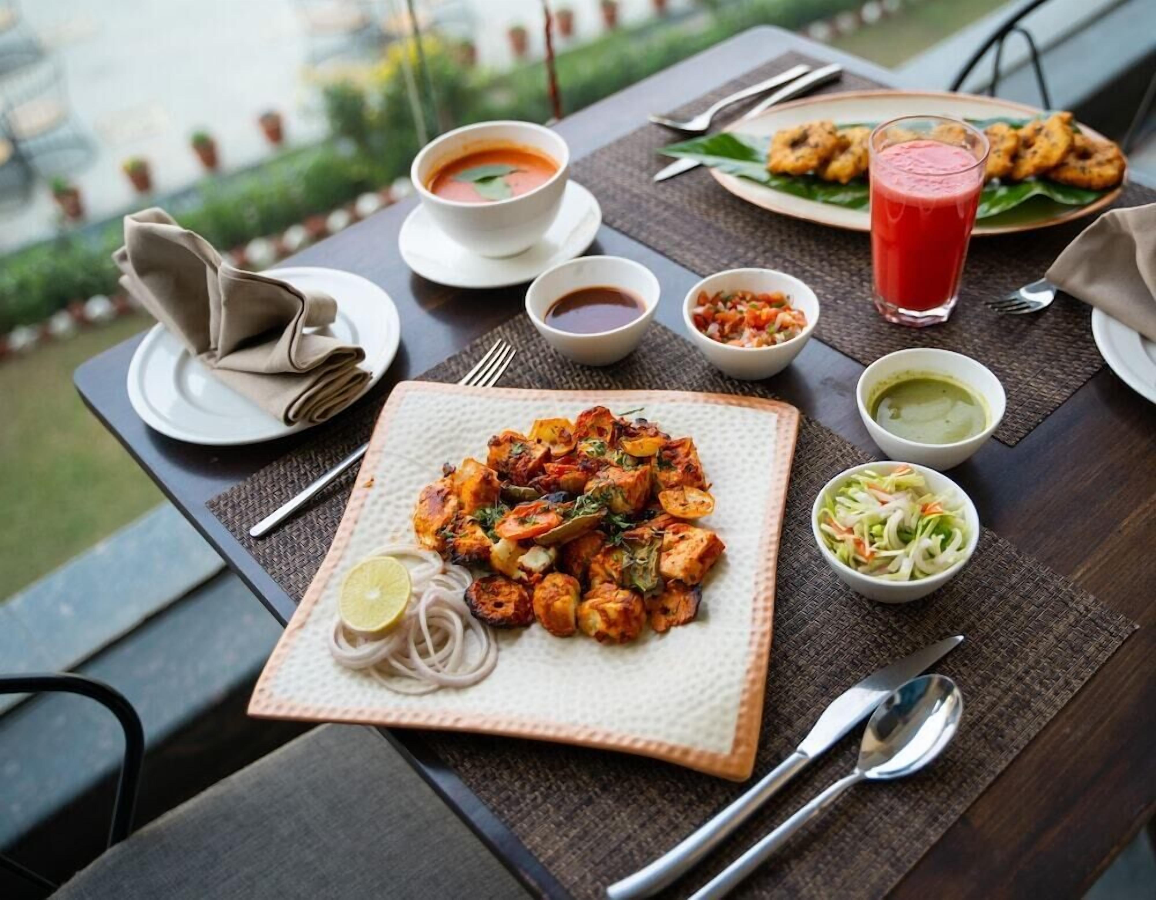 A dining table with a variety of dishes including a plate of tandoori chicken, a bowl of soup, a glass of red drink, and several side dishes.