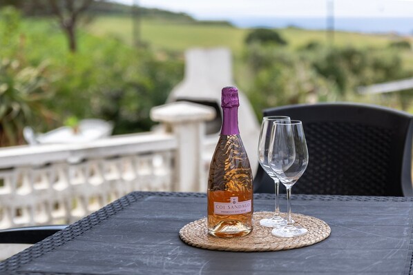 A bottle of wine and a glass on a table with a view of greenery and a white railing.