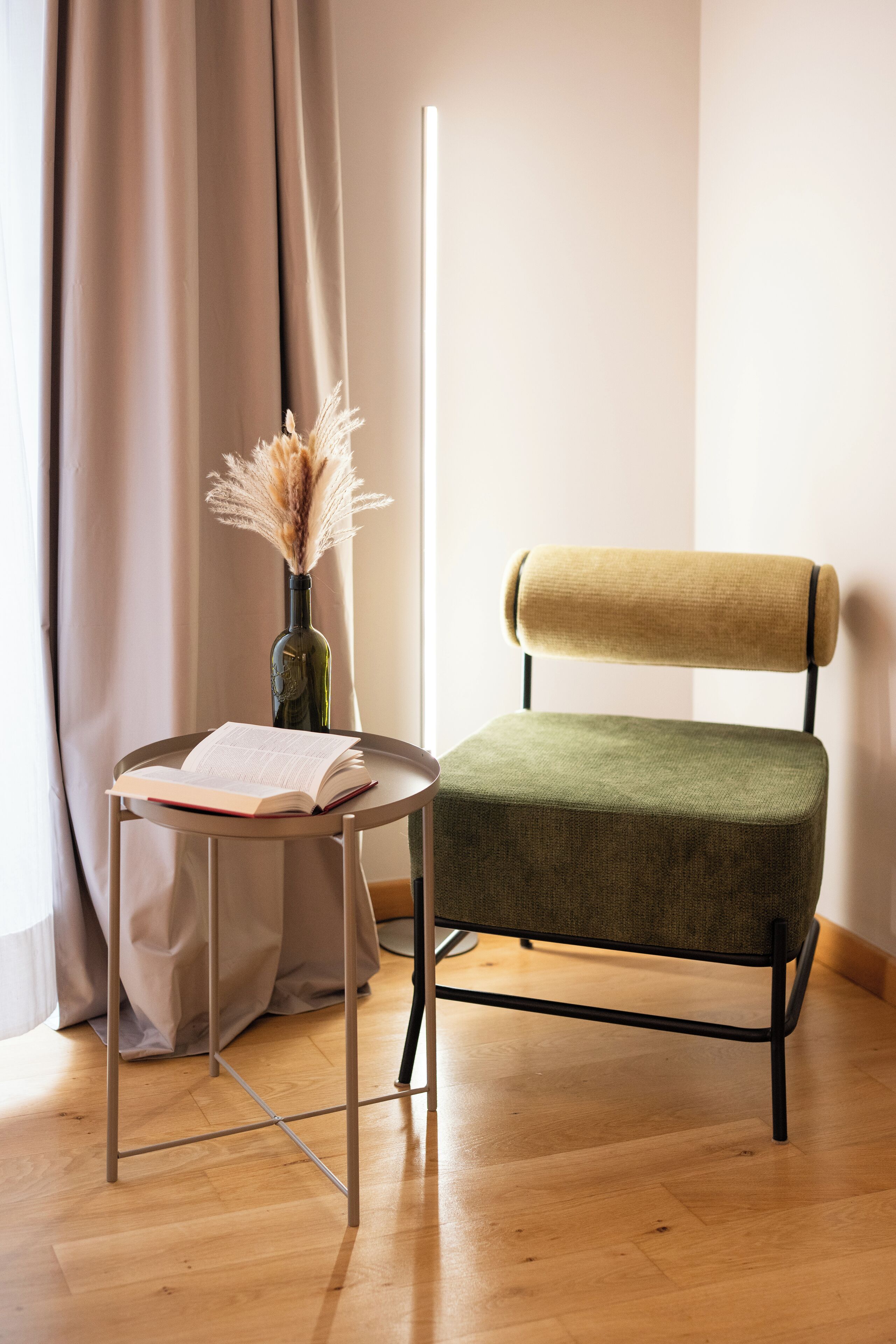 A room with a wooden floor, a green upholstered chair, a small round table with a book, and a vase with dried flowers.